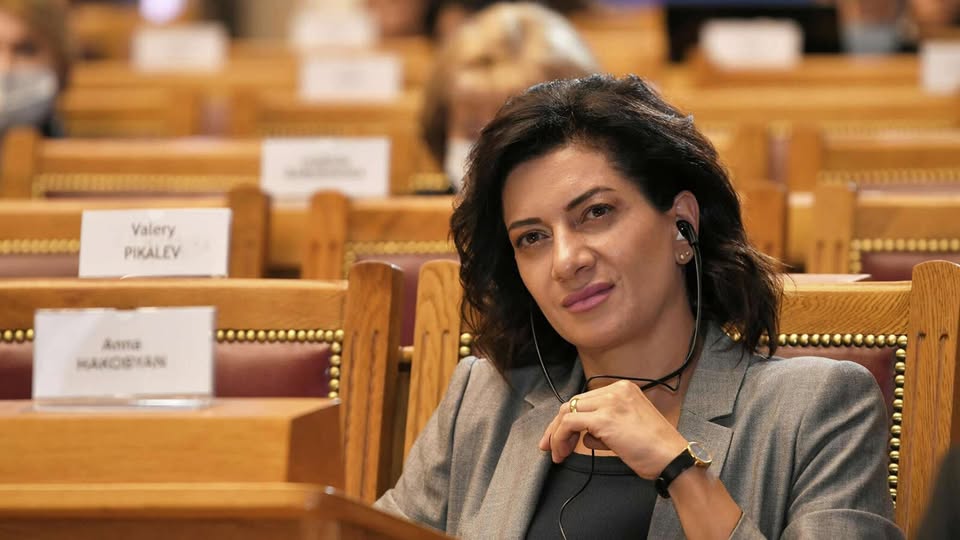 Анна Акопян в деловом костюме на фоне флагов Армении и США